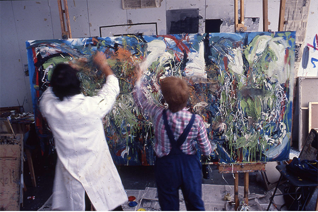 Two students in an art school painting on a large landscape oriented canvas using expressive brushstrokes.