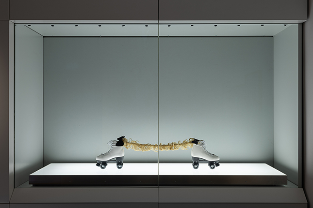 Museum glass cabinet containing two white skates on opposite ends, connected by a blonde hair extension and illuminated by a light box from below.