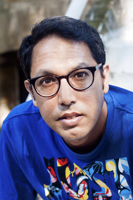 Portrait of poet Rishi Dastidar with short black hair, wearing dark framed glasses and a bright blue top with colourful illustrations on the front.