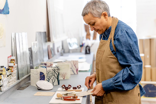 Artist and poet Suman Gujral wearing a light brown apron printmaking in a studio surrounded by printmaking supplies like linocutters and inks.