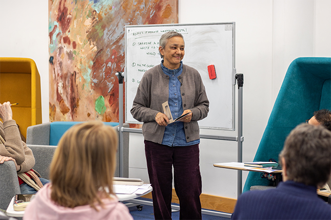 Artist and poet Suman Gujral standing in a seminar room holding a poetry book, smiling in front of a large abstract painting and whiteboard behind her. Students are sitting in front of her.