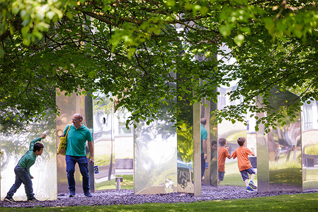 A parent and young children playing in a large maze-like mirrored sculpture surrounded by trees and grass.