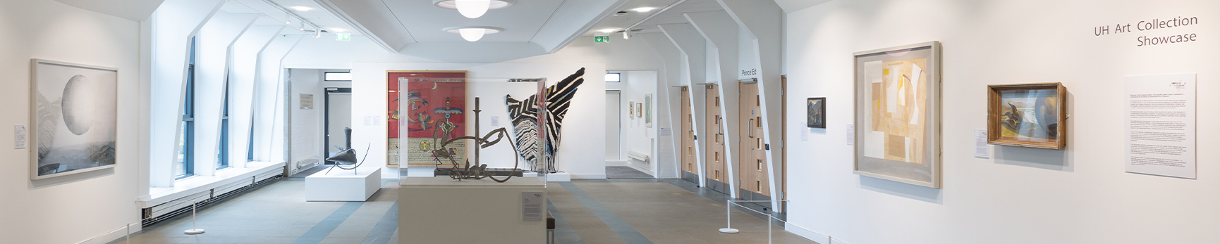 Photograph of the Chapman Gallery with a number of abstract and geometric wall-based artworks on the white walls. A sculpture on a plinth sits in the centre and is surrounded by brown benches.