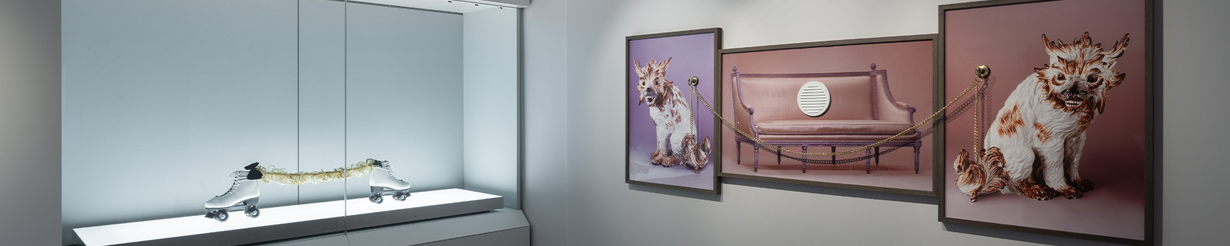 A glass museum cabinet on the left containing an assemblage of found objects including two white skates and a string of a blonde wig, illuminated by a light box. On the right are three pink and lilac coloured photographic collages containing a pink sofa and two ceramic dragon-like animals. 