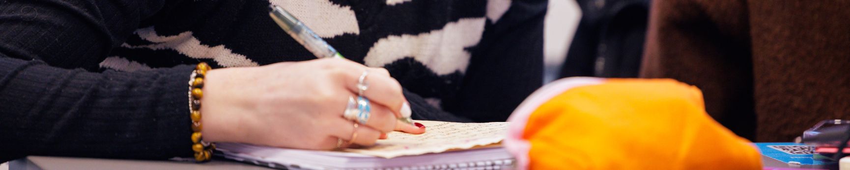 Student wearing lots of rings and bracelets holding a pen and writing words on a sheet of paper on a desk.