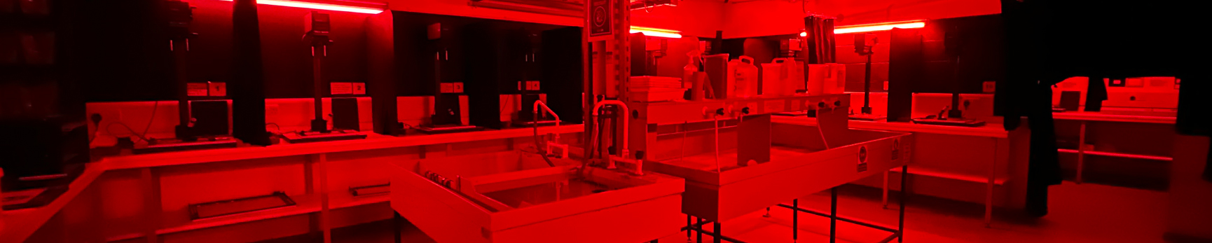 Photograph of a photographic darkroom under a red safelight with enlargers around the edges of the room and photographic chemical baths and sinks in the middle.