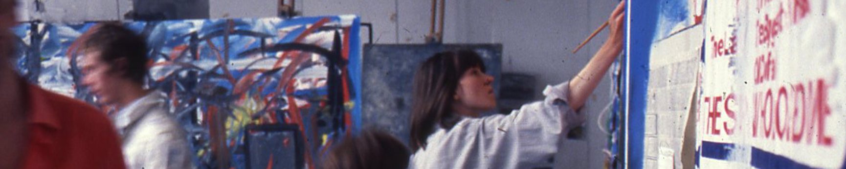 Young woman with short dark brown hair painting in an art school surrounded by abstract paintings and other students