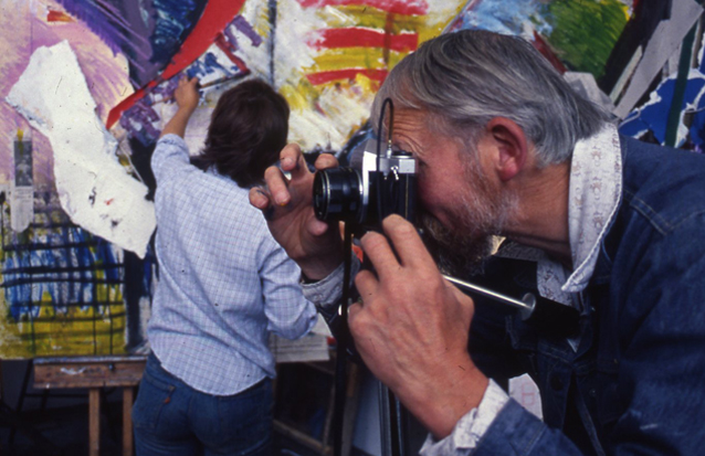 Man with grey hair taking a photo on a black film camera in front of a person painting on a large colourful surface in the background