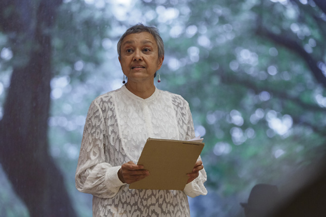 Award-winning artist and poet Suman Gujral with short grey hair and wearing a white patterned dress, reading from a brown clipboard in front of a picture of trees.