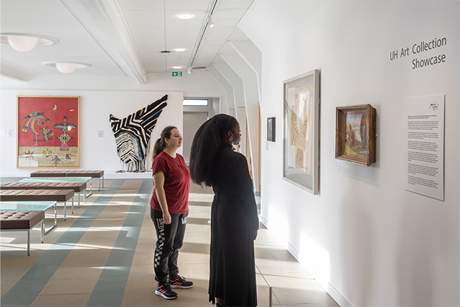 Two students looking at wall-based artworks in a white-walled gallery. There are black vinyl letters on the wall that read UH Art Collection Showcase.